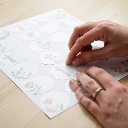 Gros plan réaliste sur des étiquettes autocollantes de baptême rondes sur une table en bois clair, avec des motifs floraux pastel et des mains qui les lissent délicatement, lumière naturelle et profondeur de champ faible.