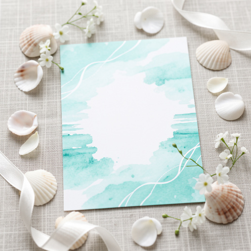 Faire-part de mariage bleu turquoise et blanc posé sur un tissu en lin, avec ruban, coquillages et fleurs blanches, ambiance lumineuse et océanique, sans texte.